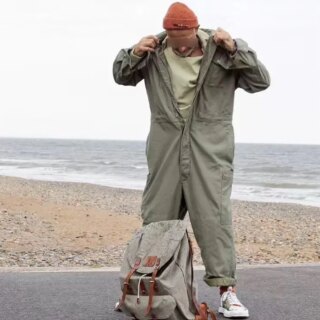 Photo d'un homme portant une combinaison de travail large style cargo avec un bonnet orange, il se tient debout devant une plage avec un sac à dos à ses pieds